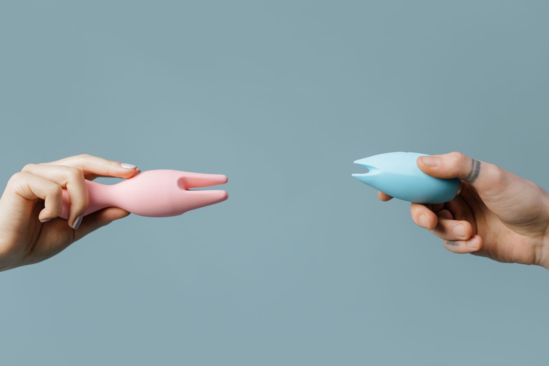 Two hands holding pink and blue silicone vibrators side by side against a pastel background.