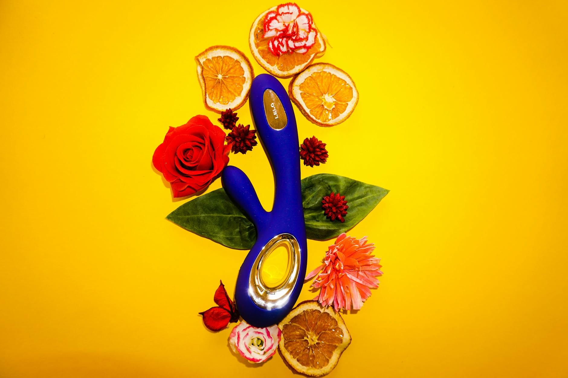 Un vibrador azul rodeado de flores y rodajas de naranja sobre fondo amarillo.